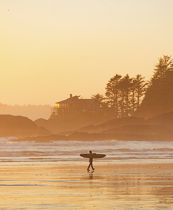 Surfer walking along Chesterman Beach at sunset with the McDiarmid family cabin in the background
