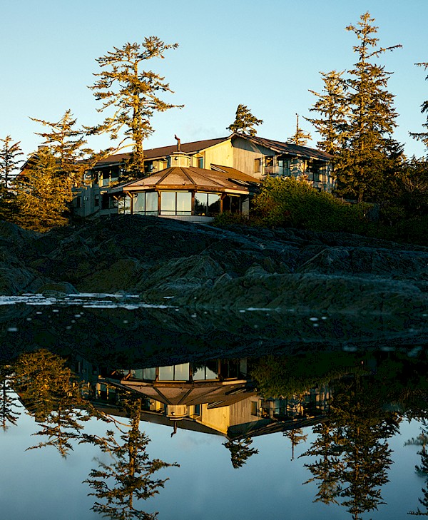 The Wickaninnish Inn reflected in a tide pool at sunrise, surrounded by coastal trees.
