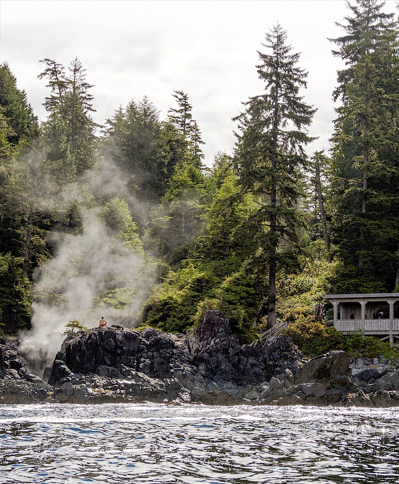 Scenic view of a steamy Hot Springs Cove with dense trees, a person sitting on a rocky outcrop, and a gazebo nestled among the forest.