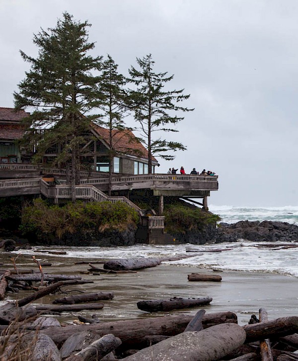 People gather on the balcony of a large wooden lodge overlooking a rocky beach with driftwood and turbulent waves, surrounded by tall trees.