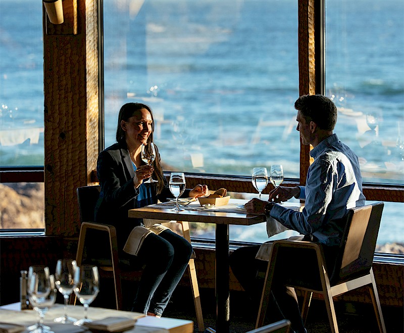 Two guests enjoying wine and oceanfront dining at a table overlooking waves through large windows