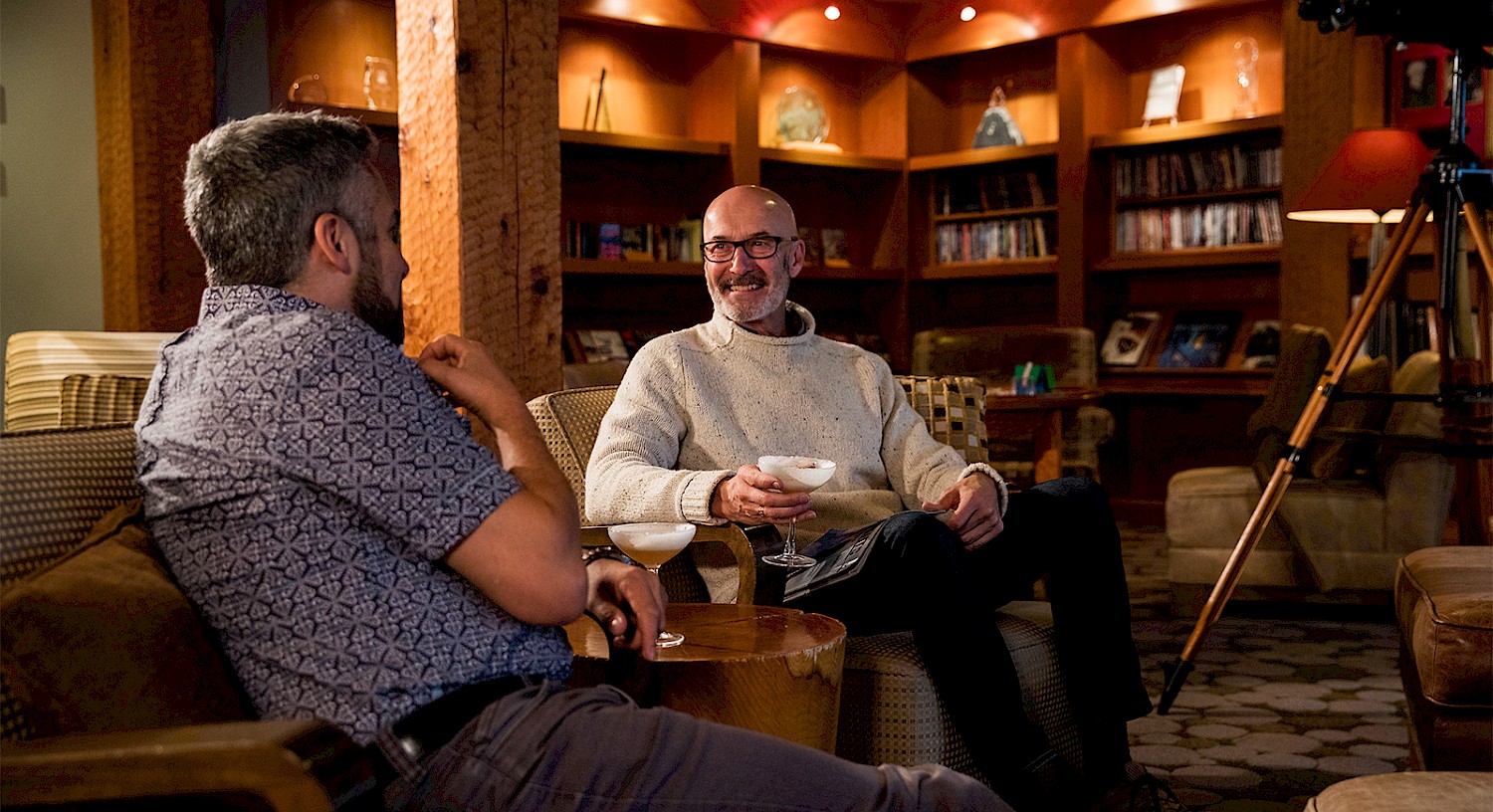 Two guests relaxing with cocktails in a warm library lounge with bookshelves and soft lighting