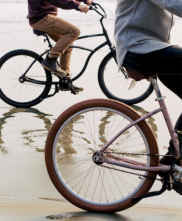 Two people riding beach cruiser bicycles along the wet sand, with reflections beneath the wheels
