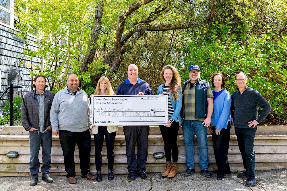 Group of eight adults standing outdoors, smiling and holding a large ceremonial check from West Coast Sustainable Tourism Association