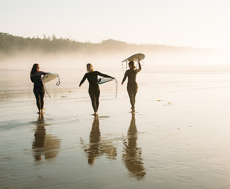 Three summer surfers holding their boards, wearing wetsuits and walking on Chesterman Beach with their reflections visible on the wet sand