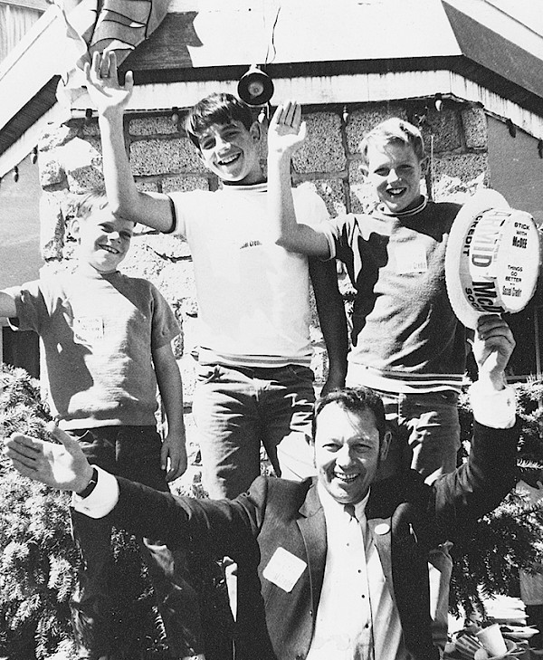 Historic black and white photo of three children and one adult waving happily outside a building. They are all smiling and appear joyful.