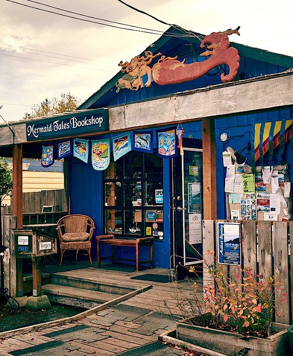 Exterior view of Mermaid Tales Bookshop featuring wooden rustic design with decor related to ocean themes, a mermaid sculpture atop the facade, and colorful banners above the window