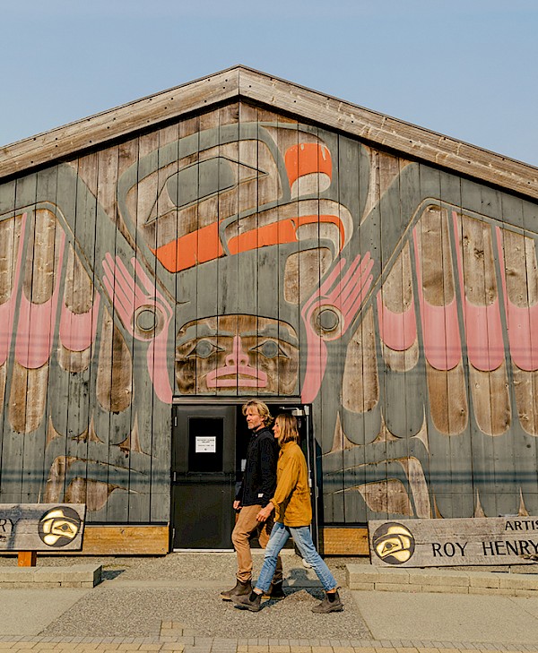 Two people walk past the Roy Henry Vickers Gallery longhouse, which features a large carving of an Indigenous art design on its wooden facade.