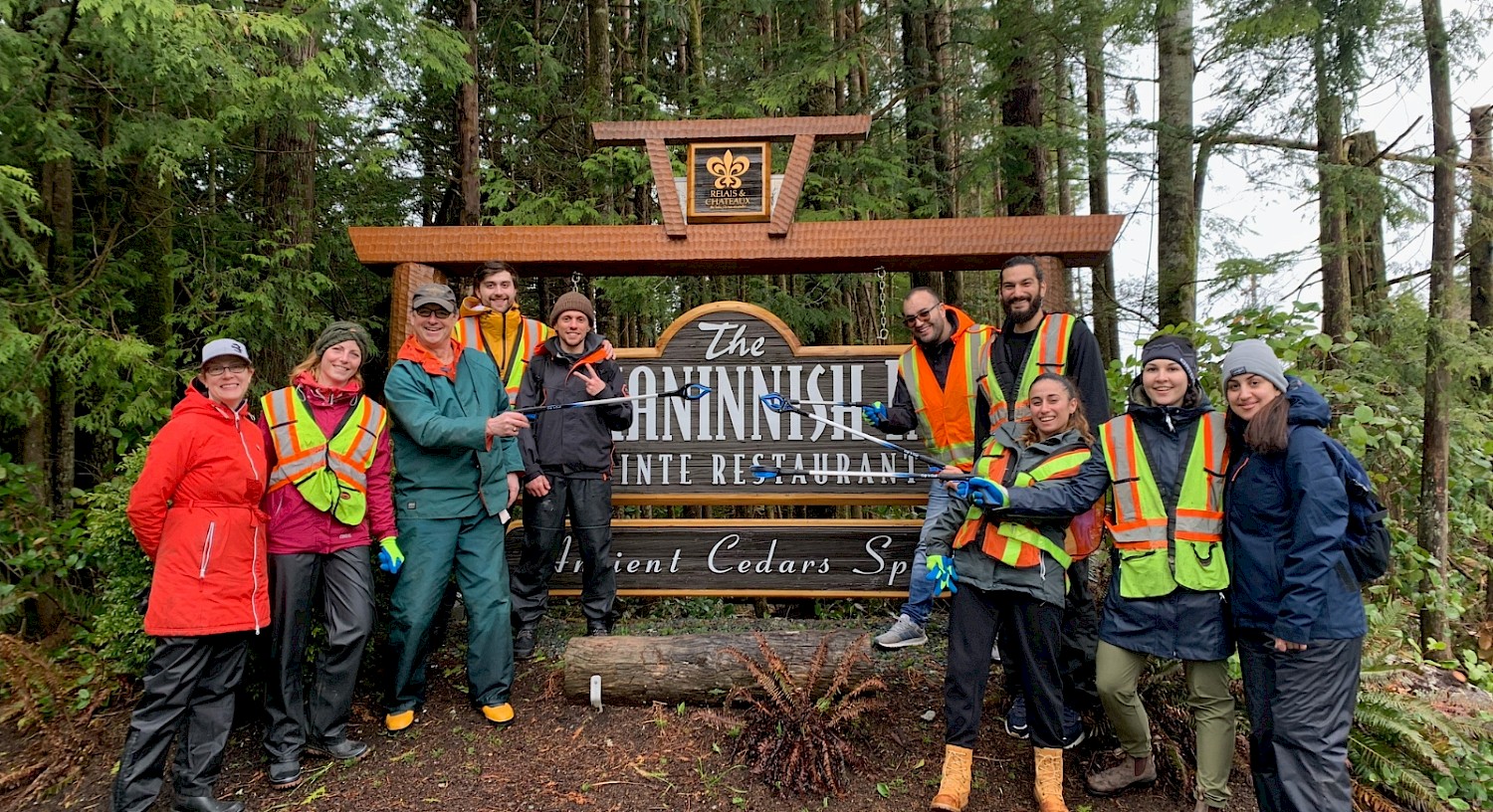 Group of team members posed in front of the roadside Wickaninnish Inn sign wearing safety vests, holding grabber tools, with a forested backdrop