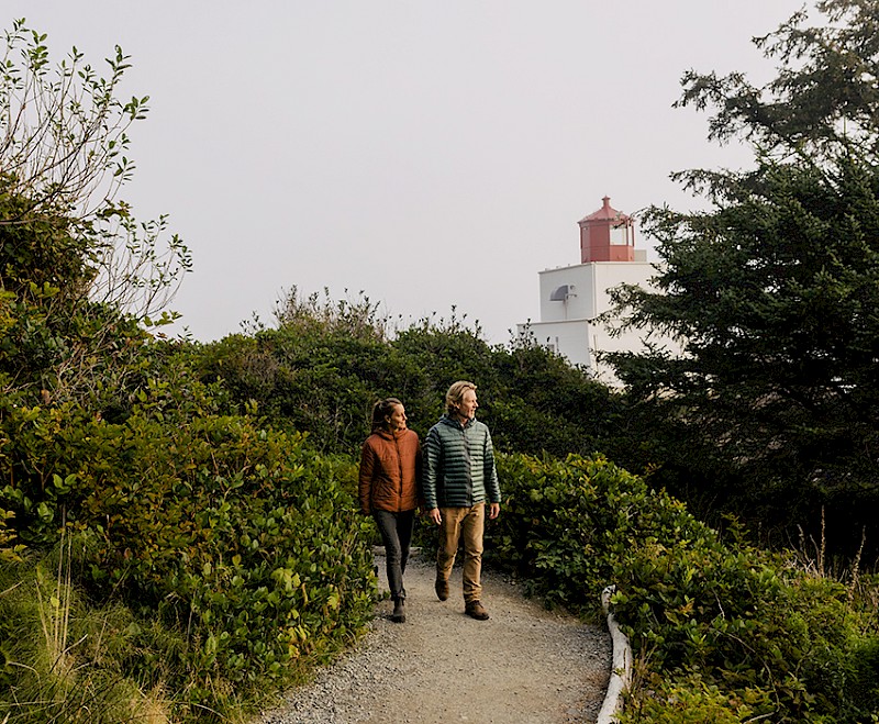 Couple walking a forested section of the Wild Pacific Trail with Amphitrite Point Lighthouse in the background