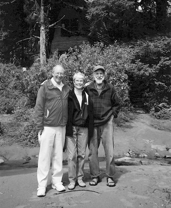 Howard and Lynn McDiarmid standing with carver Henry Nolla on a beach, surrounded by coastal forest