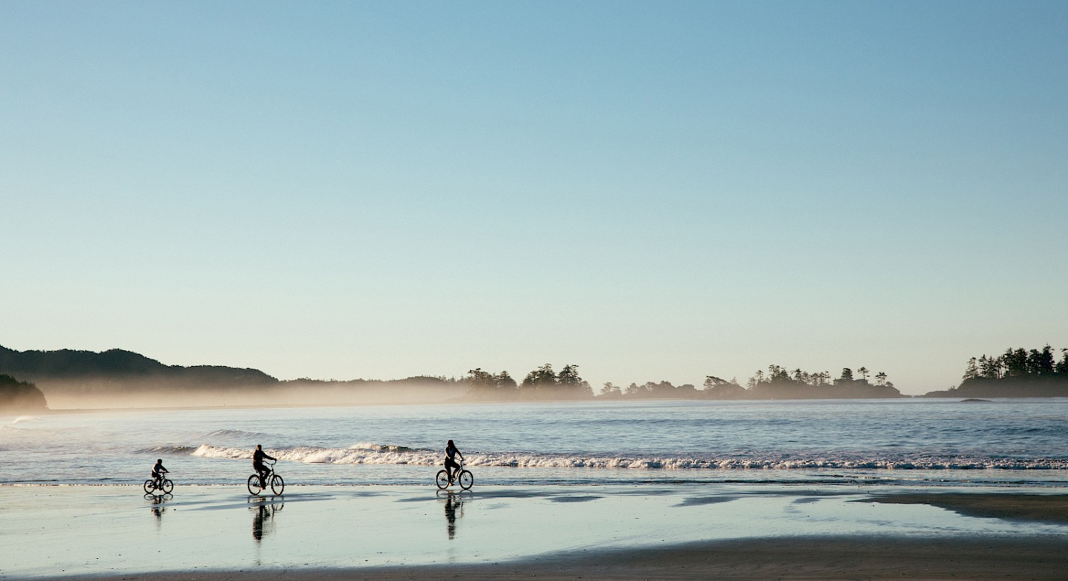 Three people ride bicycles along a tranquil Chesterman Beach with a clear blue sky. Waves gently reach the shore, and Frank Island is visible in the background.