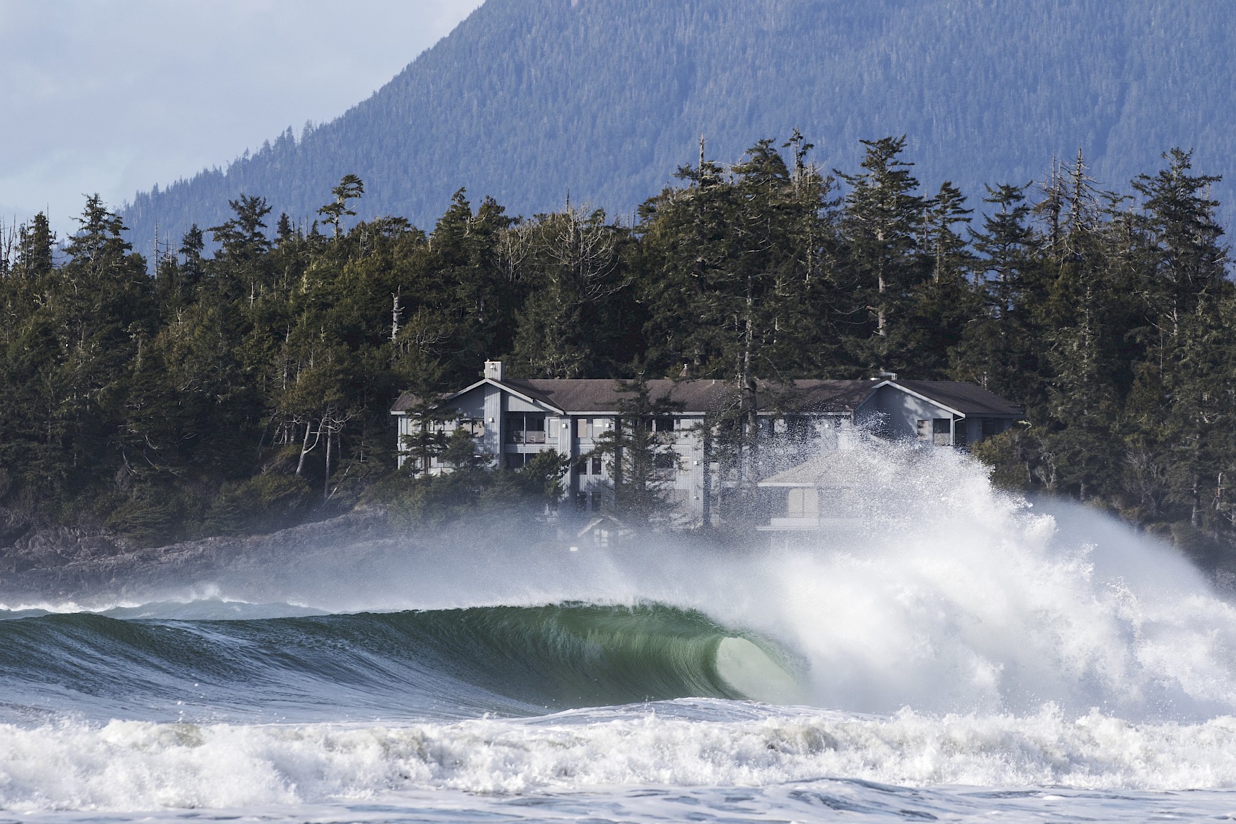 A large wave crashes ashore with the Inn, forest, and mountains in the background